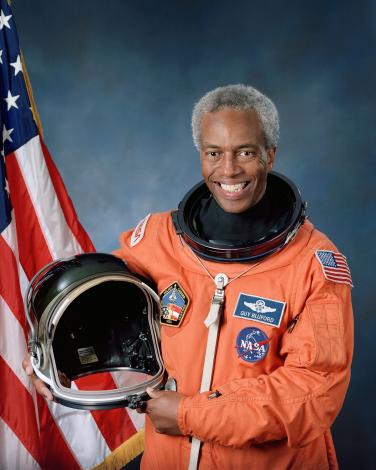 Guion Bluford's official NASA photo. He wears an orange suit and holds his helmet. The American flag is on the left side (to the viewer) of the photo and Bluford is smiling.