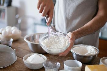 A baker is whisking a bowl of flour. Other bowls of ingredients are on the table in front of them.