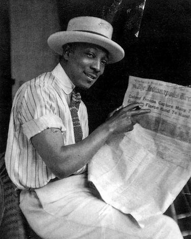 Portrait of James Van Der Zee. He wears a showboater hat, a short sleeved shirt and tie, slacks, and is holding a cigar and a newspaper.